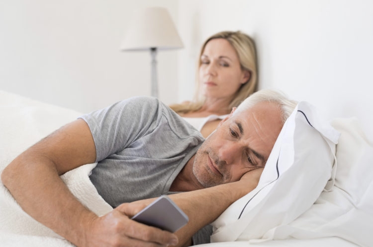 Jealous woman spying her husband mobile phone while he is reading a message. Senior couple in bed while wife is angry as husband using smartphone. Senior husband ignoring wife and texting on smartphone.